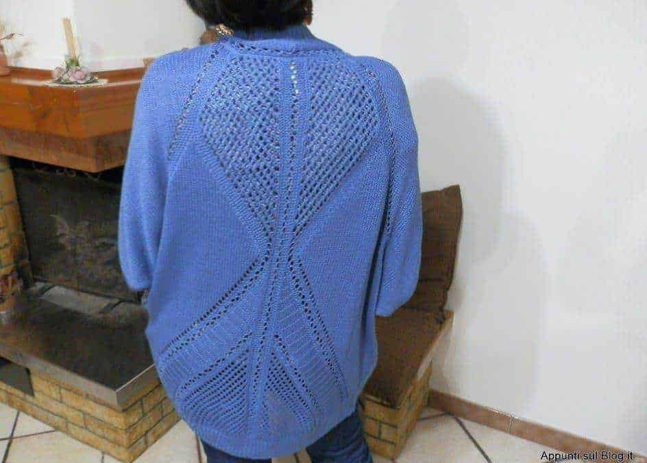 Lei, indossando un maglione blu con dettagli traforati, si trova in un ambiente domestico con un camino in mattoni sullo sfondo.