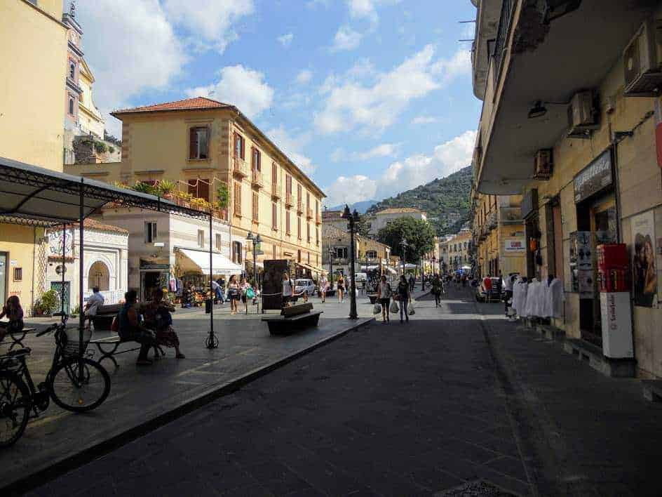 Vicolo pedonale nel centro storico di Maiori, un caratteristico paese italiano con edificio antichi, negozi, bar e persone che passeggiano sotto un cielo sereno e nuvoloso, con colline sullo sfondo.