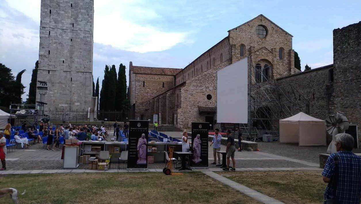 Cinema all'aperto in una storica piazza con antiche mura e torre, partecipanti che aspettano l'inizio della proiezione sotto un cielo nuvoloso, spazio di eventi culturali e sociali.