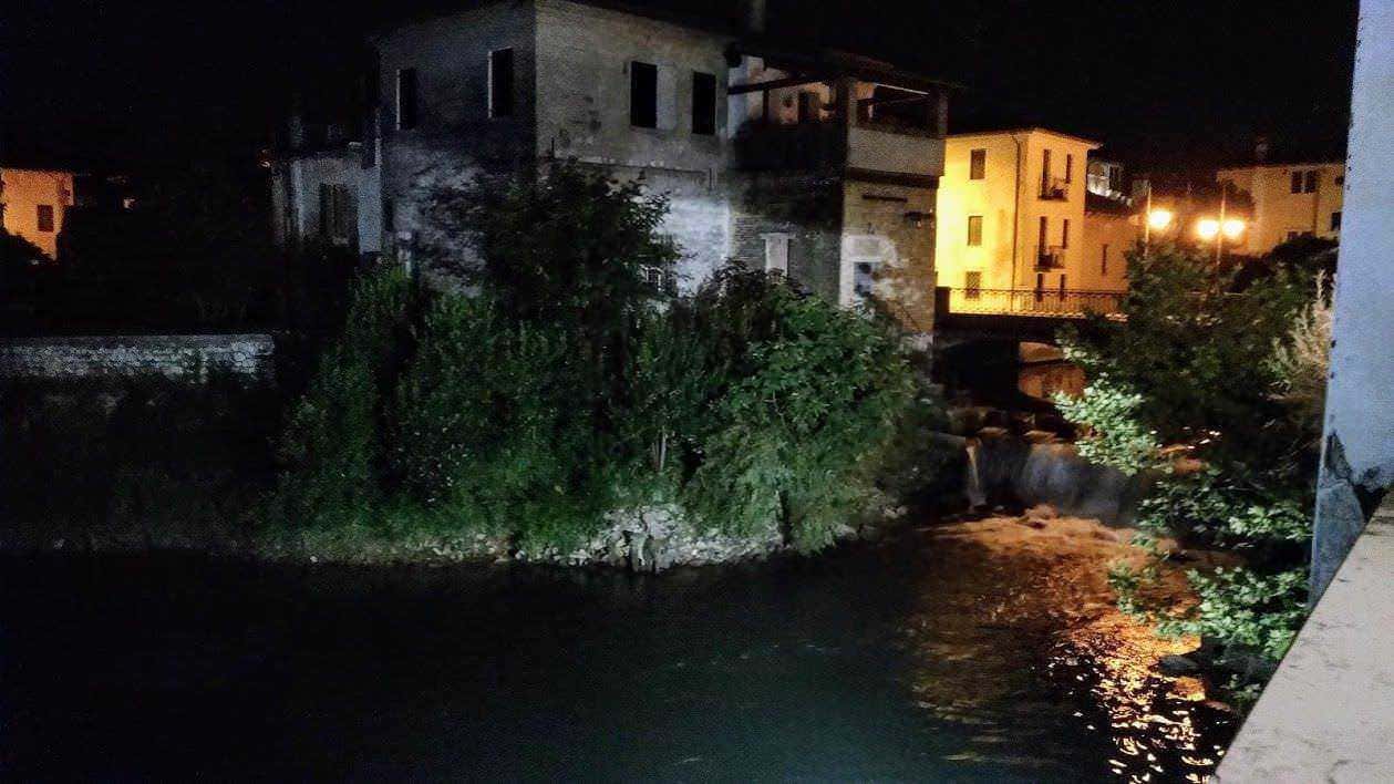 Tratto di fiume urbano di notte, illuminato da luci calde, con edifici antichi sullo sfondo e vegetazione lungo le rive, rappresenta un paesaggio suggestivo tipico di un ambiente cittadino.