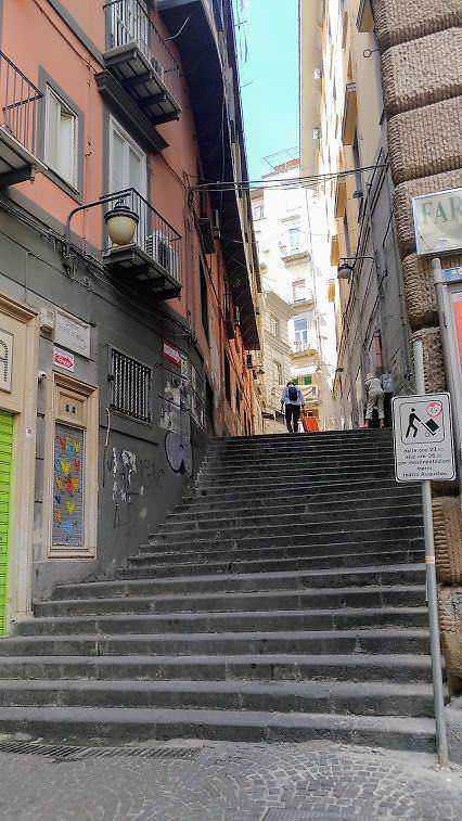 Coppie di scale in zona urbana di Napoli con edifici storici, strada stretta e ringhiere di ferro, rappresentando un tipico scorcio cittadino.
