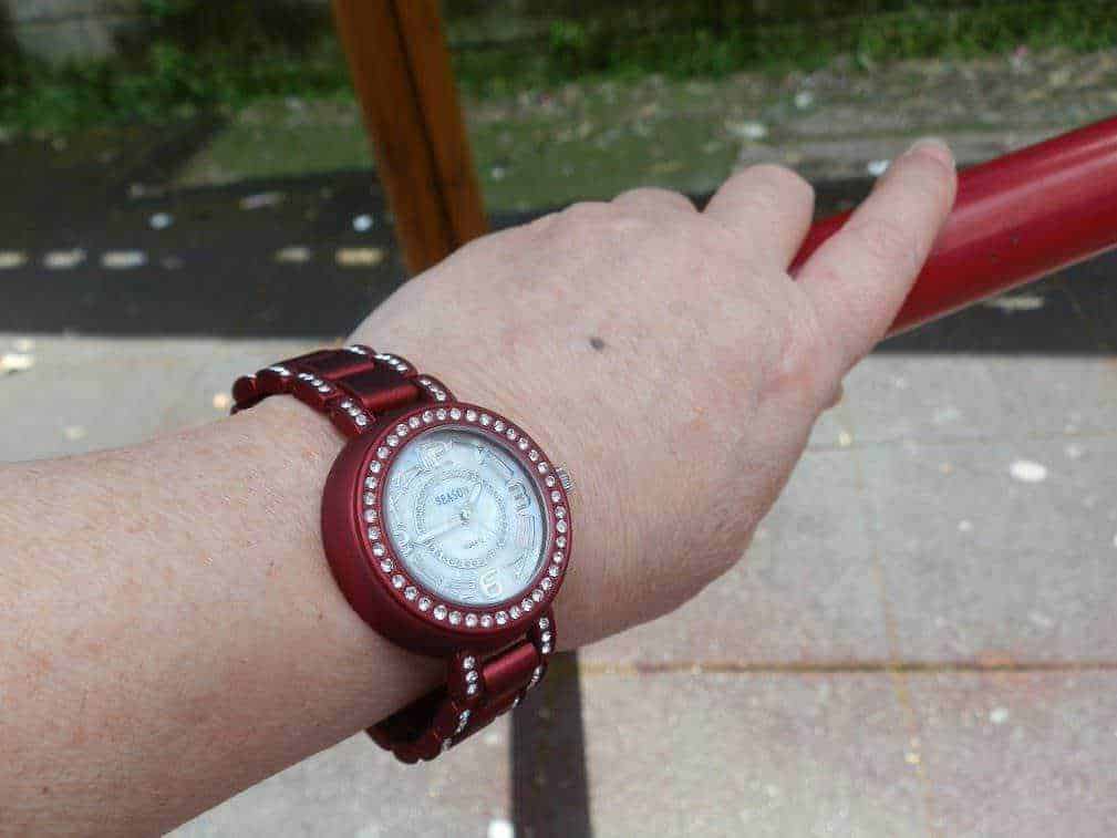 Bracciale e orologio rosso con pietre di cristallo, indossato su un polso femminile, vista all'aperto su passeggiata urbana, accessori eleganti e alla moda.