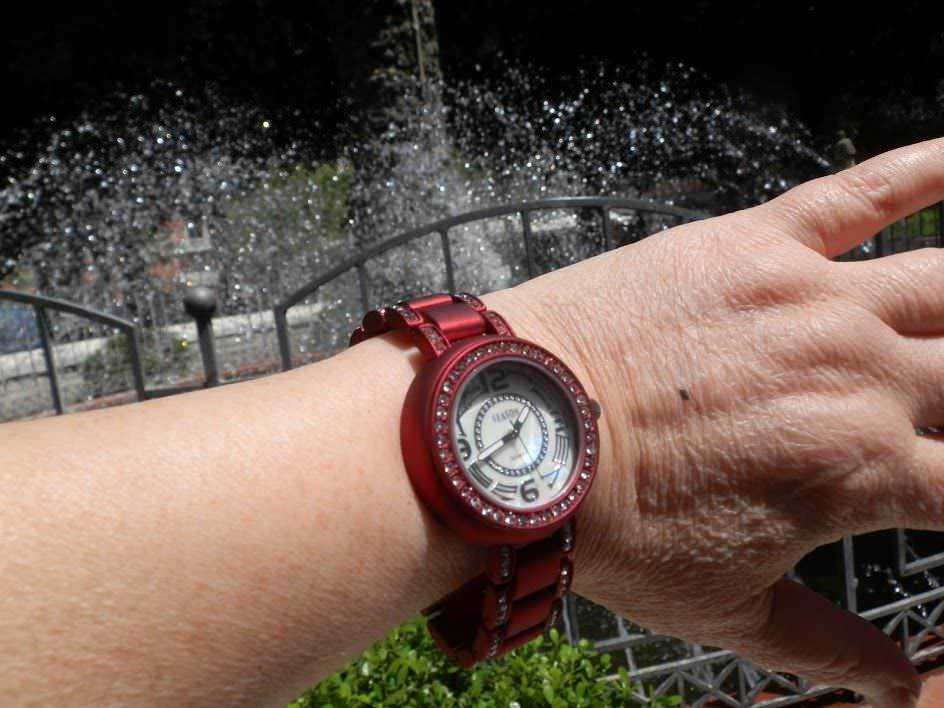 Orologio da donna rosso con cinturino in metallo e cinturino, con pietre colorate e quadrante bianco, indossato su un polso di donna con sfondo di fontana e giardino.