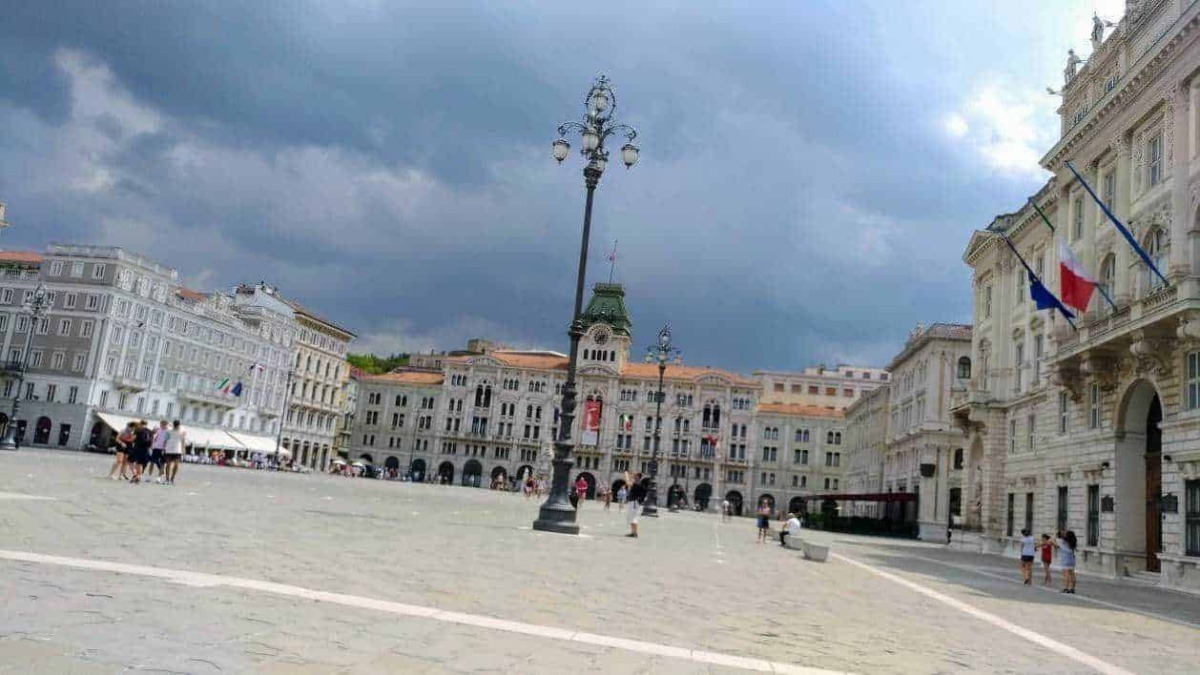 Luminoso piazza storica con edifici in stile classico e bandiere italiane, sotto un cielo nuvoloso, zona centrale di una città italiana.