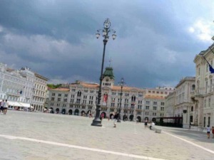 Luminoso piazza storica con edifici in stile classico e bandiere italiane, sotto un cielo nuvoloso, zona centrale di una città italiana.