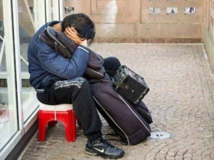 Invecchiamento precoce: Aspetto di un giovane senza lavoro e senza soldi seduto su una sedia rossa all'esterno di un negozio, con bagagli e radio portatile, simbolo di disagio sociale e povertà.