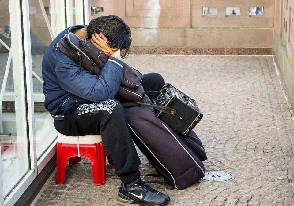 Invecchiamento precoce: Aspetto di un giovane senza lavoro e senza soldi seduto su una sedia rossa all'esterno di un negozio, con bagagli e radio portatile, simbolo di disagio sociale e povertà.