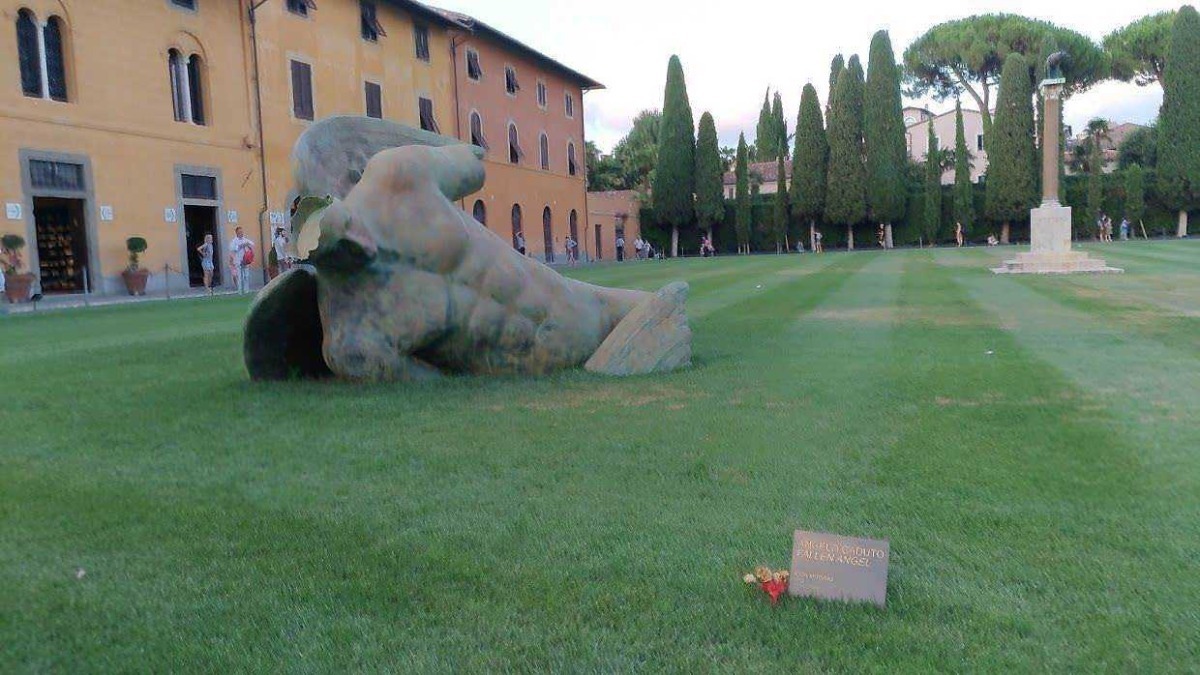 Scultura moderna di un volto umano in bronzo nel parco con alberi e monumento commemorativo, area verde ben curata e visitatori in background, arte pubblica e cultura urbana.