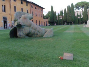 Scultura moderna di un volto umano in bronzo nel parco con alberi e monumento commemorativo, area verde ben curata e visitatori in background, arte pubblica e cultura urbana.