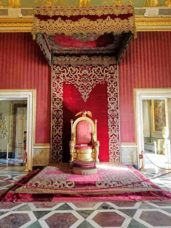 Regina nel trono dorato in uno stile barocco, decorato con ricami e velluto rosso, all’interno di un palazzo storico con pareti elaborate e pavimento a scacchi.