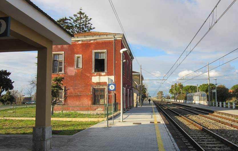 Edificio storico rosso lungo la stazione ferroviaria sul binario, con piattaforma pedonale, linee elettriche aeree e paesaggio naturale circostante, ideal per foto di viaggio e cultura locale.