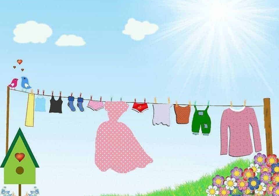 Vestiti stesi ad asciugare al sole in un cortile con prato verde e fiori colorati, scenario di una giornata di primavera o estate, con nuvole e cielo azzurro.