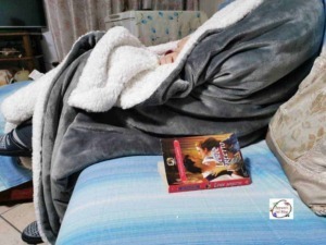Sognatore sdraiato su un divano con coperta grigia e libro aperto, ambiente casalingo confortevole, atmosfera rilassante, ideale per leggere o riposare.