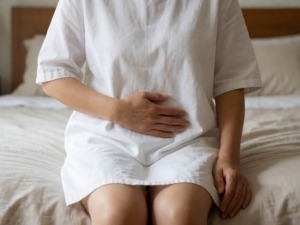 Dolore addominale e ginecologico, donna seduta su letto con mano sulla pancia, problema di salute femminile, disfunzione o malattia ginecologica, immagine medica per blog di salute e medicina.