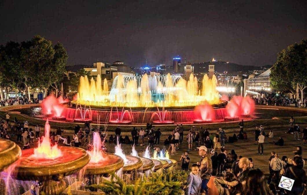 Colorata fontana a illuminazione notturna in un centro città affollato, con luci vivaci e molte persone che si intrattengono e passeggiano, creando un’atmosfera di festa e divertimento.