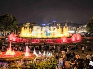 Colorata fontana a illuminazione notturna in un centro città affollato, con luci vivaci e molte persone che si intrattengono e passeggiano, creando un’atmosfera di festa e divertimento.