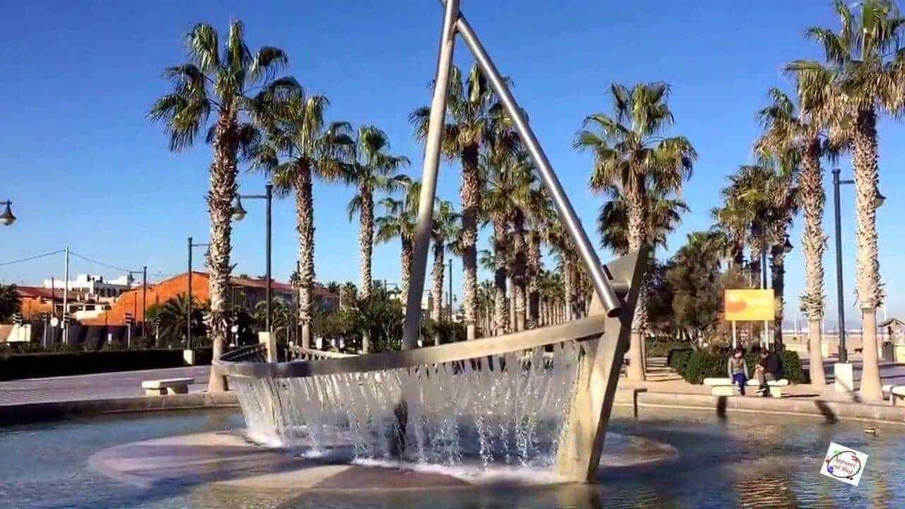 Fioriera moderna in metallo con fontana di acqua al centro, palme e area pedonale in背景 in una città costiera soleggiata, perfetta per rilassarsi e passeggiate.