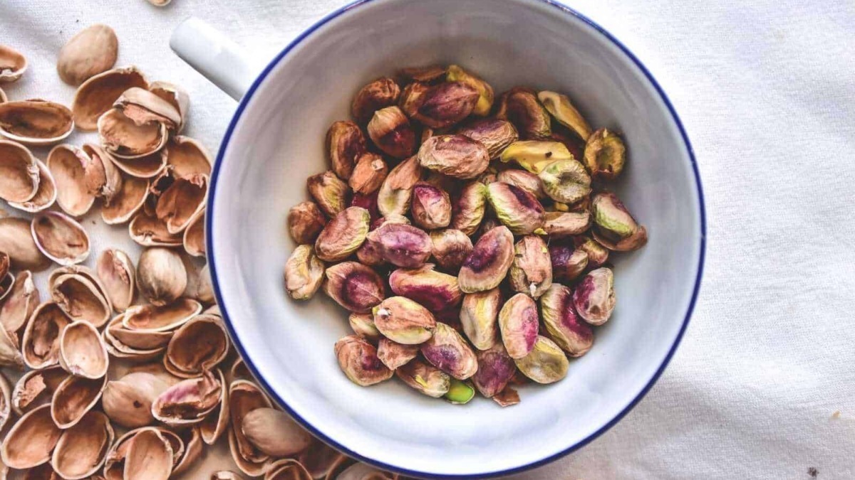 Semi di pistacchio in una ciotola bianca con bordo blu, circondati da gusci di pistacchio aperti, su tovaglia chiara, ideale per ricette e snack salutari.