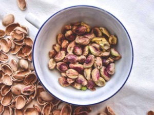 Semi di pistacchio in una ciotola bianca con bordo blu, circondati da gusci di pistacchio aperti, su tovaglia chiara, ideale per ricette e snack salutari.