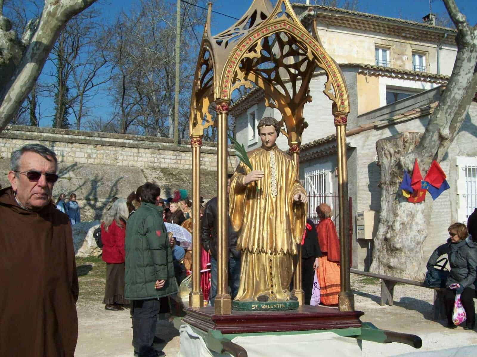 Calco in legno di San Valentino durante una festa religiosa all'aperto con partecipanti e decorazioni, in un cortile di paese con case storiche e alberi nudi, giornata di sole.