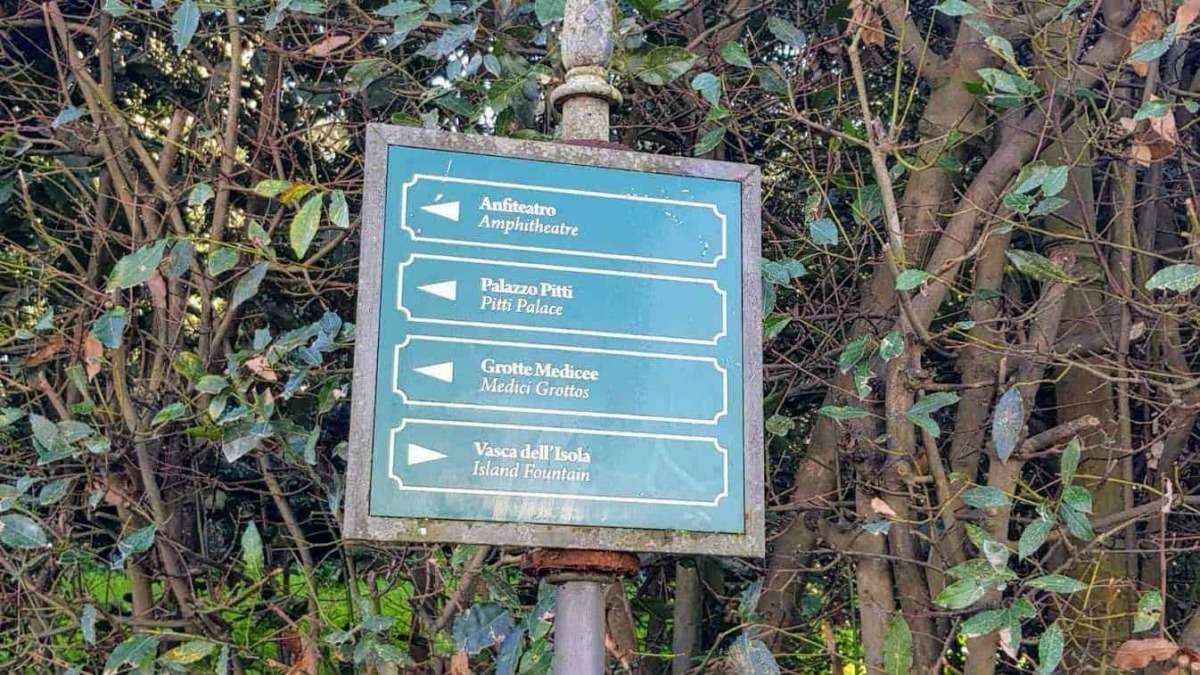 Parcheggio stradale con cartello indicatore di direzioni in stile vintage, tra arbusti e vegetazione, mostra i nomi delle principali attrazioni turistiche di un'area storica.