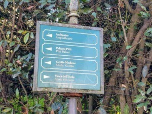 Parcheggio stradale con cartello indicatore di direzioni in stile vintage, tra arbusti e vegetazione, mostra i nomi delle principali attrazioni turistiche di un'area storica.