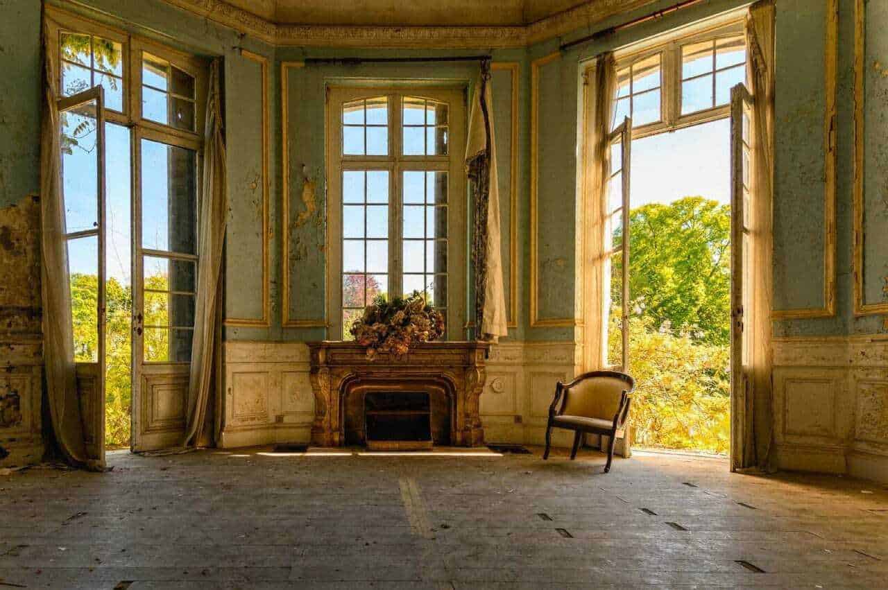 Ancora di una stanza di lusso abbandonata, con grandi finestre che lasciano entrare luce naturale, pareti decorate e un camino antico con fiori, in un ambiente ricco di storia e decadimento.