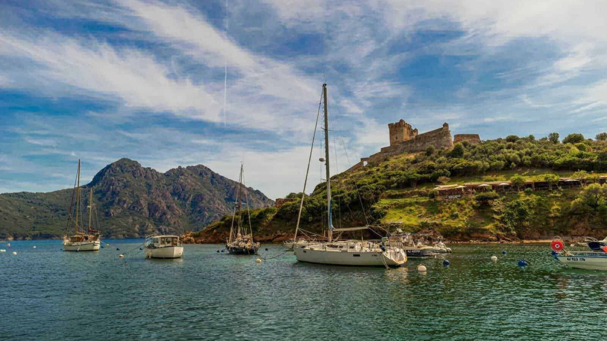 Barche a vela ormeggiate nel lago con il castello in rovina sullo sfondo, in un paesaggio naturale con montagne e cielo azzurro.