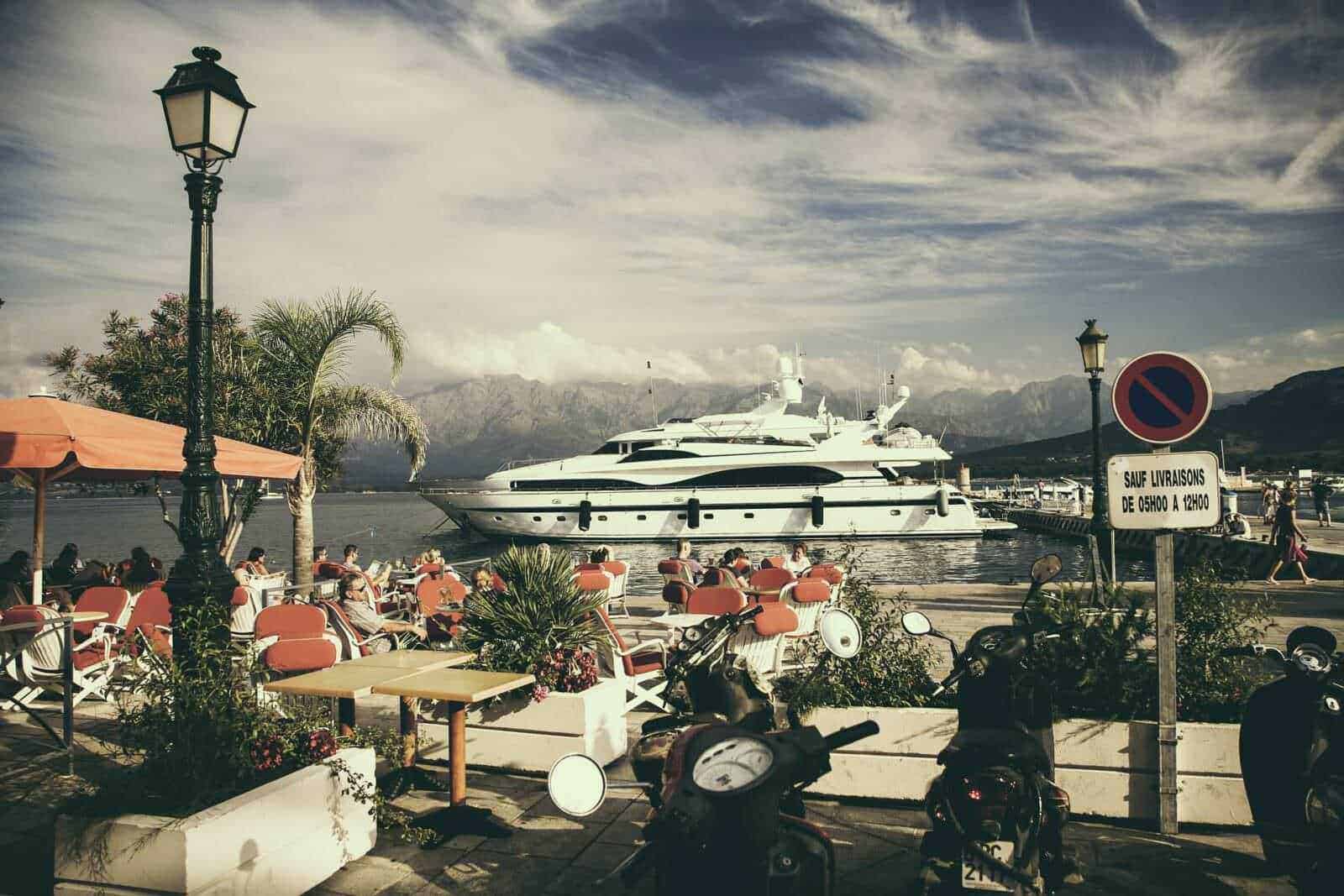 Come arrivare in Corsica da Roma, info viaggi 2 Faro di pietra lungo il porto con barche di lusso e motoscooter, atmosfera rilassante con persone sedute ai tavoli, montagne sullo sfondo, cielo nuvoloso, segnale di divieto di sosta.
