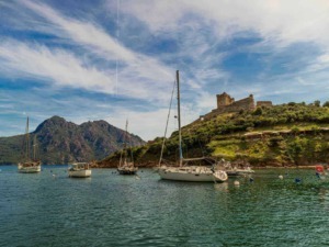 Barche a vela ormeggiate nel lago con il castello in rovina sullo sfondo, in un paesaggio naturale con montagne e cielo azzurro.