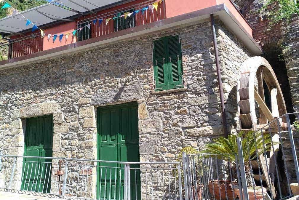 Vecchia casa in pietra con ruota idraulica e finestre con persiane verdi, situata in un paesaggio montano, decorata con bandierine colorate, atmosfera rustica e storico locale.
