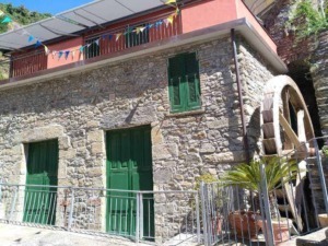 Vecchia casa in pietra con ruota idraulica e finestre con persiane verdi, situata in un paesaggio montano, decorata con bandierine colorate, atmosfera rustica e storico locale.