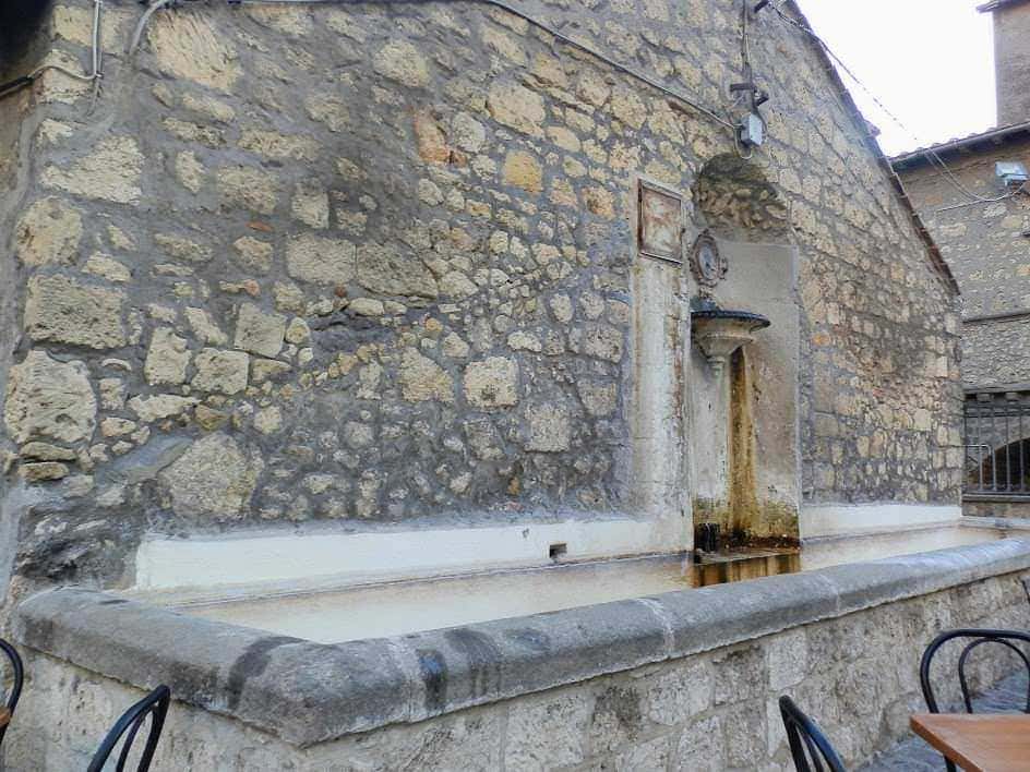 Vecchio fontanile in pietra con abbeveratoio in marmo, situato in un borgo storico italiano, caratterizzato da pareti in pietra e archi tradizionali, ideale per fotografie di paesaggi rurali e architettura storica.