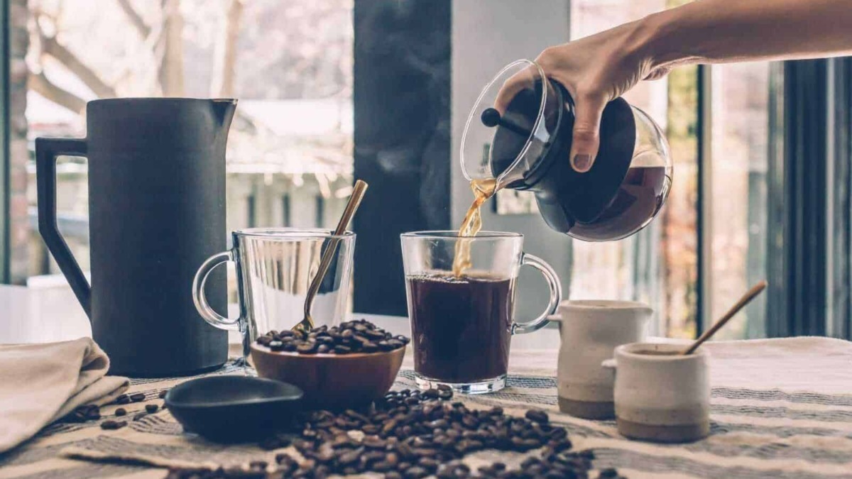 Un'illustrazione di una colazione con caffè, dove il caffè appena preparato viene versato in un bicchiere trasparente, circondato da chicchi di caffè, tazze e una brocca di caffè.