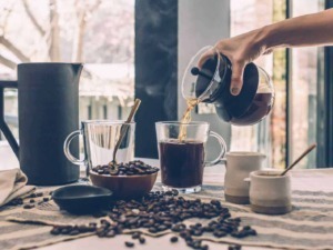 Un'illustrazione di una colazione con caffè, dove il caffè appena preparato viene versato in un bicchiere trasparente, circondato da chicchi di caffè, tazze e una brocca di caffè.