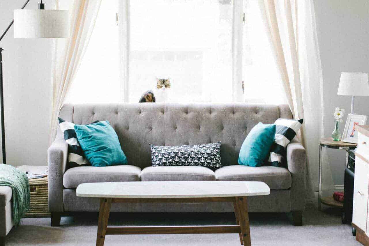 Atmosfera accogliente in soggiorno con divano tondo, cuscini blu e decorazioni moderne su tavolino e scaffale, grande finestra con tende leggere, perfetta per ambienti di stile e comfort.