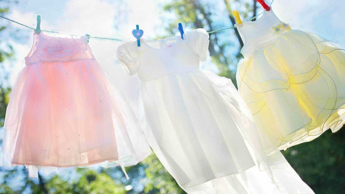 Vestiti per bambine appesi ad una corda da bucato, con clip colorate, in un giardino soleggiato con albero e cielo limpido, simbolo di primavera e giochi all'aperto.
