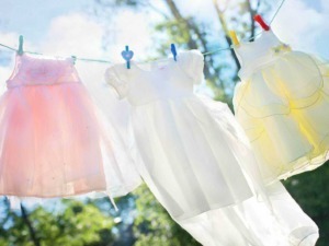 Vestiti per bambine appesi ad una corda da bucato, con clip colorate, in un giardino soleggiato con albero e cielo limpido, simbolo di primavera e giochi all'aperto.