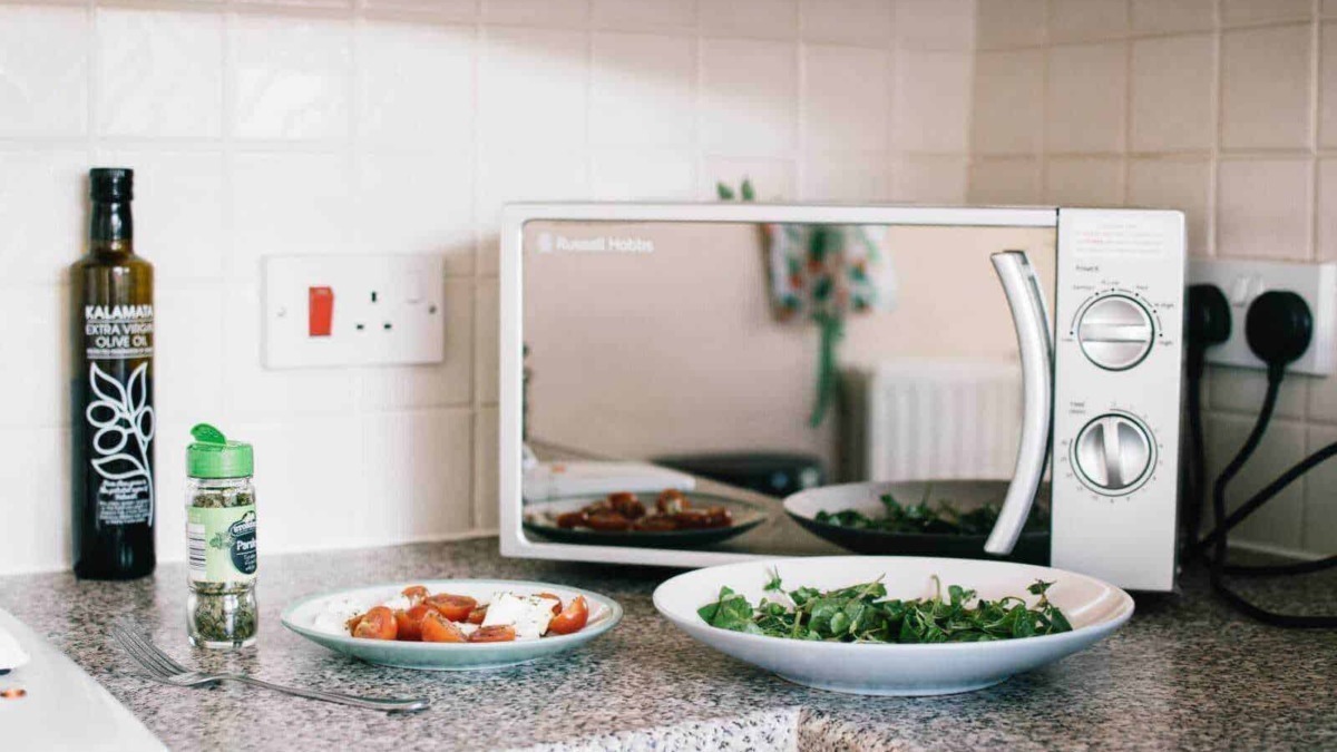 Forno a microonde con piatti di verdure fresche e pomodori sul piano di cucina, oliolare e sale, ambientazione moderna con pareti chiare.