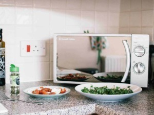 Forno a microonde con piatti di verdure fresche e pomodori sul piano di cucina, oliolare e sale, ambientazione moderna con pareti chiare.
