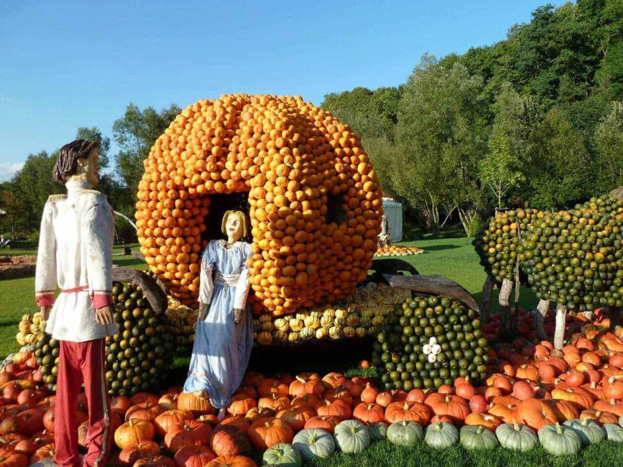 Cantiere di zucche e ortaggi decorativi in forma di carrozza, in un parco, con figure di persone in abiti tradizionali in mostra, ambiente autunnale colorato.