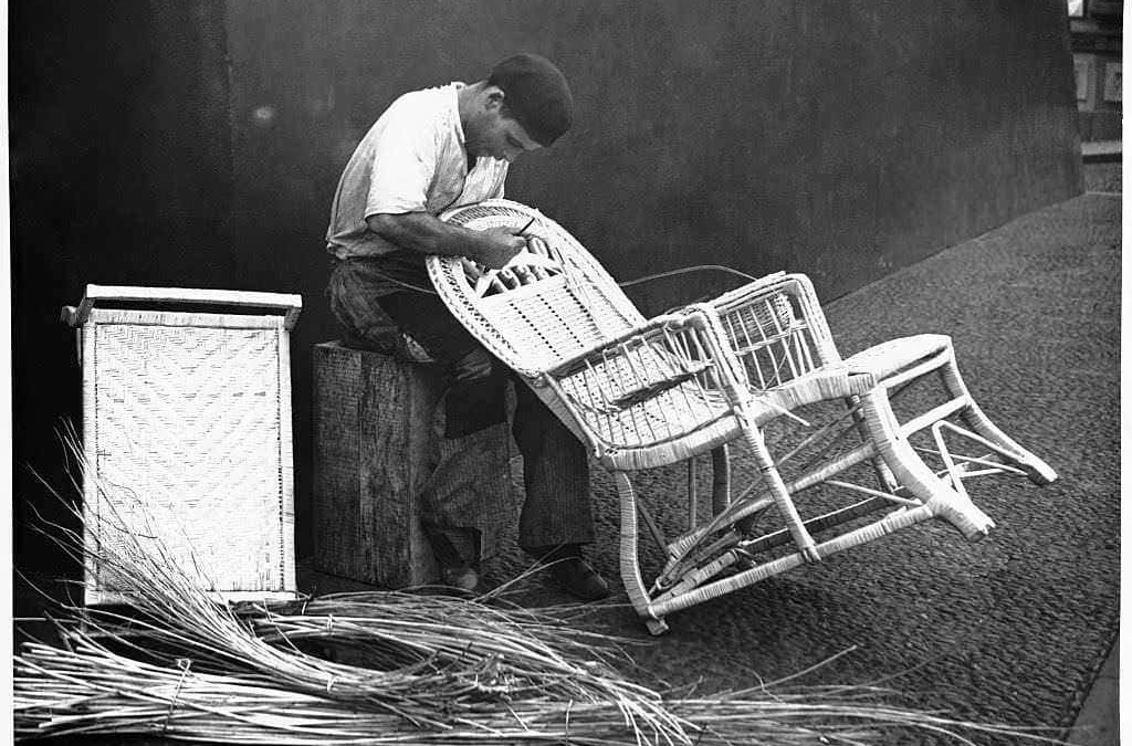 Sarto di mobili in vimini che ripara sedie di vimini, con materiali di fibra naturale sul pavimento, in un ambiente artigianale.