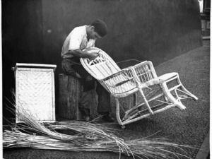 Sarto di mobili in vimini che ripara sedie di vimini, con materiali di fibra naturale sul pavimento, in un ambiente artigianale.