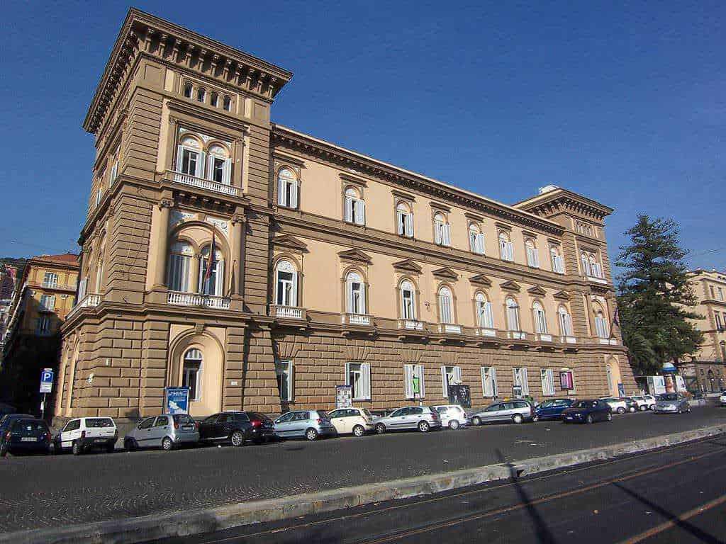 Palazzo storico in stile classico con grandi finestre e decorazioni architettoniche dettagliate, situato in una via cittadina con auto parcheggiate e cielo azzurro.