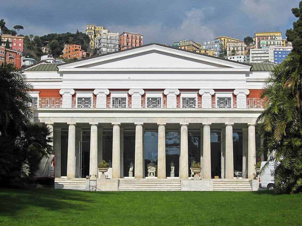 Magnifico edificio neoclassico con colonne e dettagli architettonici eleganti, immerso in un verde prato, con sfondo di case colorate su colline. Perfetto esempio di architettura storica in ambiente urbano.