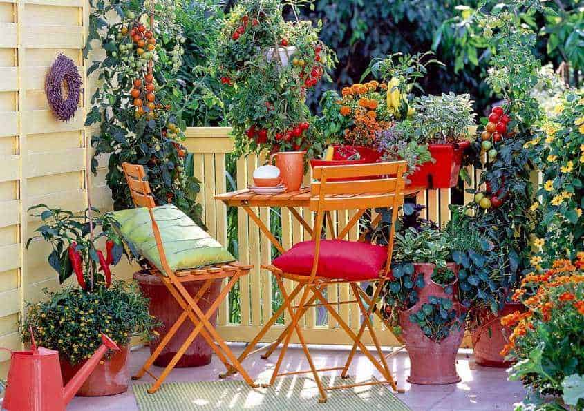 Colorato angolo caffè in terrazza con tavolino e sedie in legno circondato da piante e fiori, ideale per momenti di relax all'aperto in un giardino accogliente e fiorito.