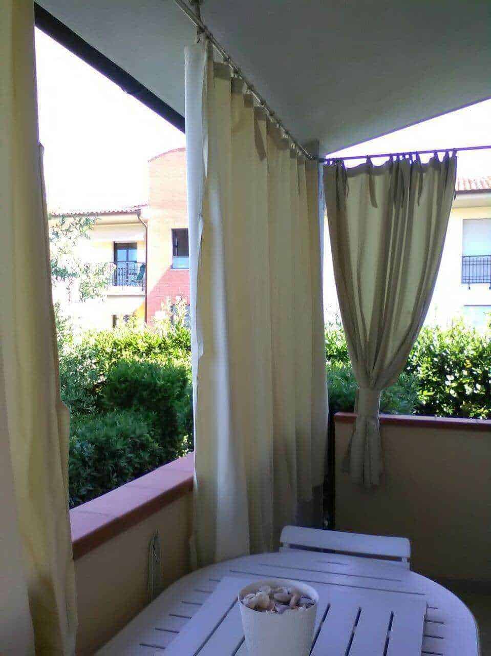 Balcone di un appartamento con tende chiare e vista su giardino e edifici residenziali, ambiente tranquillo e soleggiato, ideale per momenti di relax all'aperto in un giardino accogliente e fiorito.