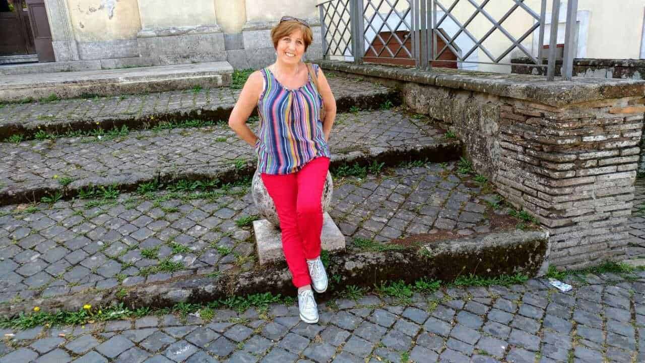 Vicina donna seduta su un sedile in pietra su un'area di ciottoli urbani con sfondo di costruzioni storiche.