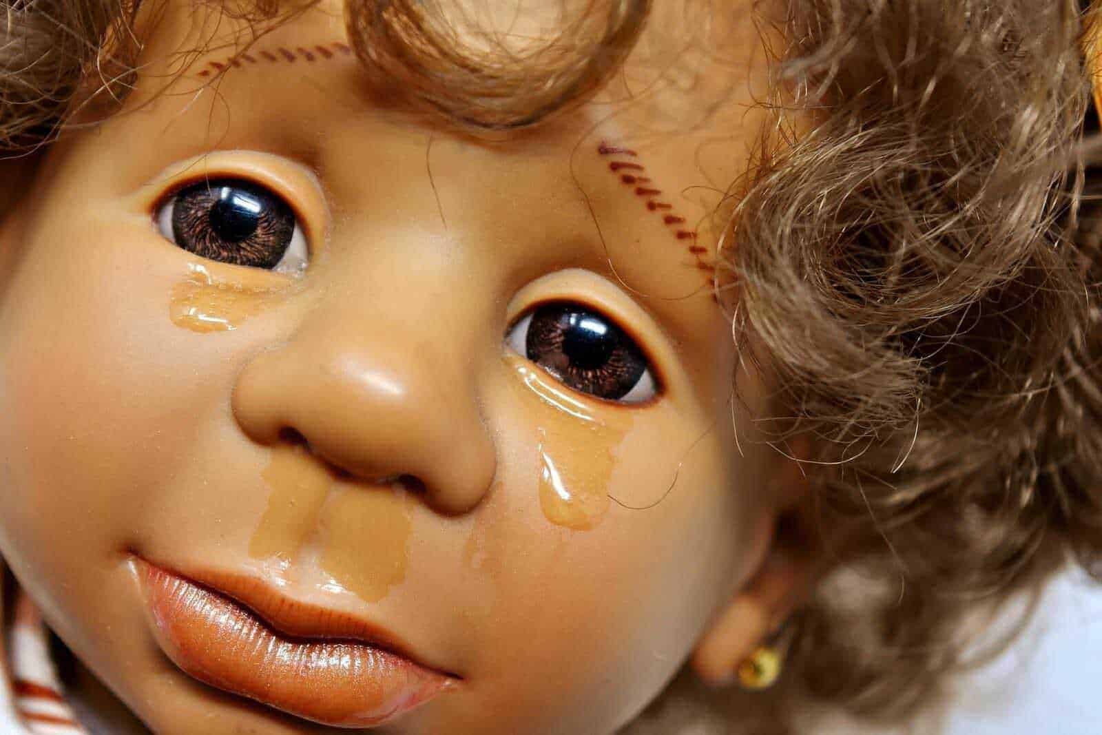 Occhi di bambola fragili con lacrime e trucco, dettagli realistici in primo piano, rappresentano emozioni intense, fotografia iperrealista con attenzione ai particolari del viso e testo su blog.