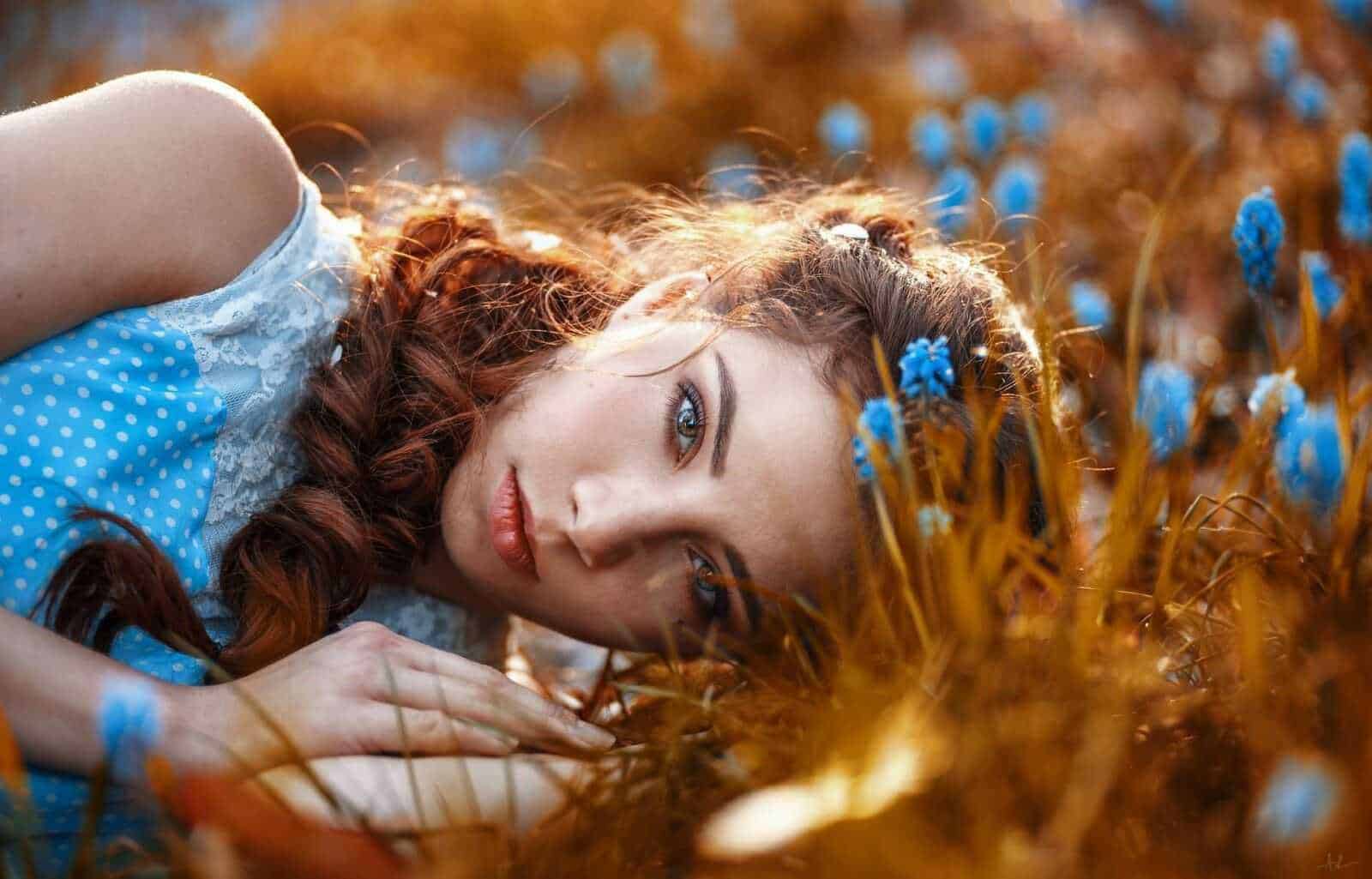 Femminile con capelli ricci e occhi azzurri che sdraiata tra fiori blu in un campo soleggiato.